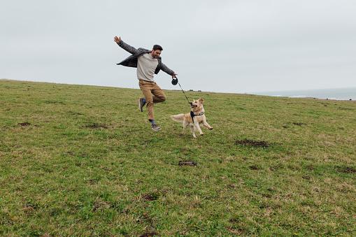 A wide angle front view of a man and his golden retriever dog frolicking and having fun on a field in Newton by the Sea. They are both having a fantastic time and the young man is jumping up in the air