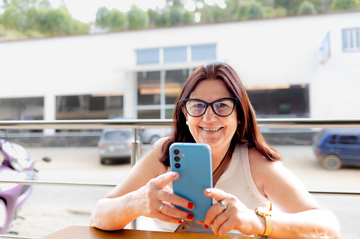Mature Brazilian woman smiling at the camera while sitting at an outdoor cafe table and using her smartphone for payments, money transfers, and social networking. Illustrating mature individuals embracing technology and financial apps
