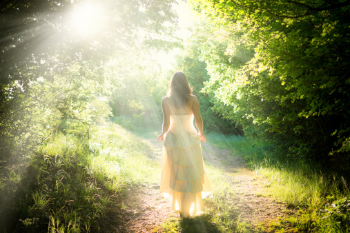 Beautiful young woman wearing elegant white dress walking on a forest path with rays of sunlight beaming through the leaves of the trees