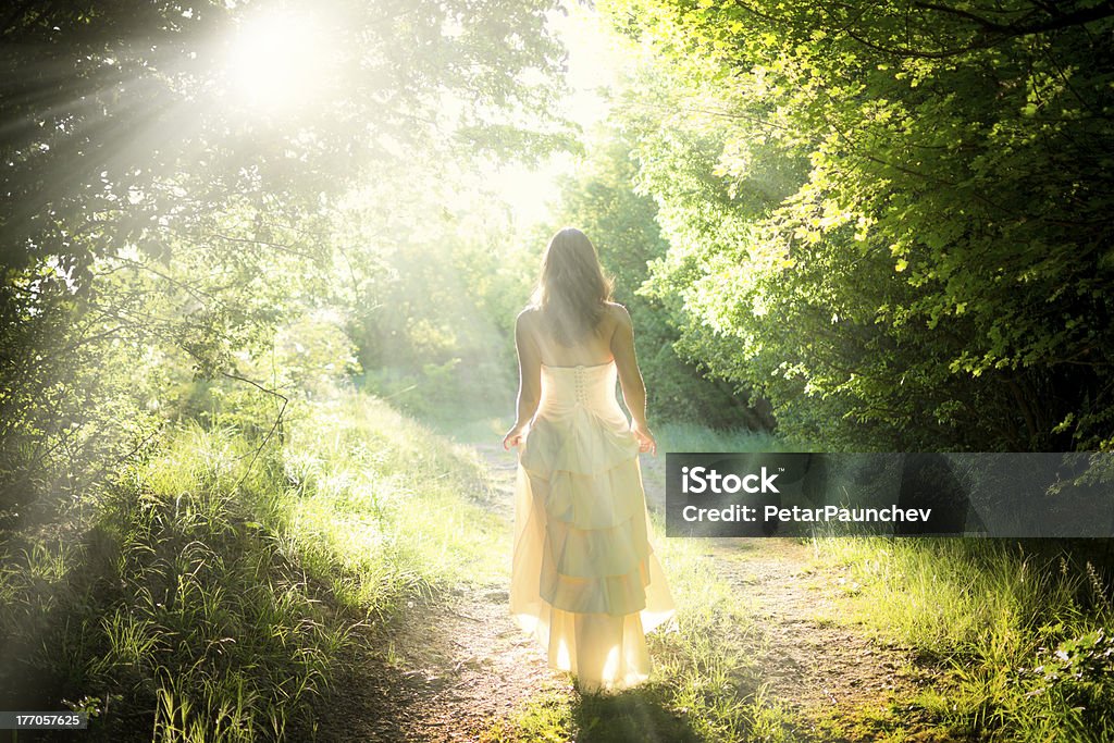 Walking fairy Beautiful young woman wearing elegant white dress walking on a forest path with rays of sunlight beaming through the leaves of the trees Women Stock Photo