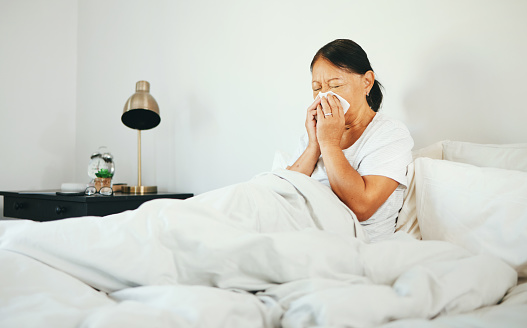 Senior woman, blowing nose and sick in bed with allergy, virus from bacteria with health and wellness at home. Sinus infection, medical condition with toilet paper for illness and sneezing in bedroom