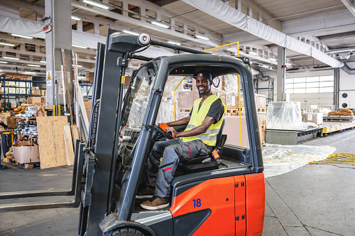 African American airport warehouse worker driving a forklift and moving aircraft cargo.