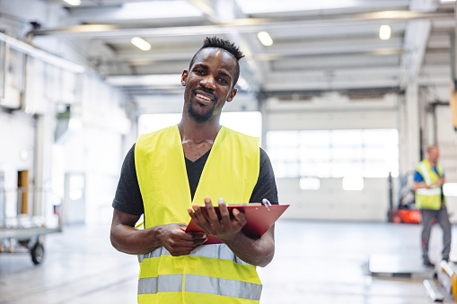 Portrait of an African American airport worker holding a notepad. He is wearing a reflective vest standing in a warehouse.