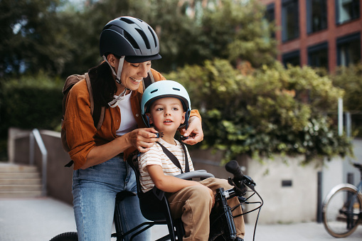 Mother fastening sons' bike helmet on head, carring him on child bike carrier or seat. Mom commuting with a young child through the city on a bicycle.
