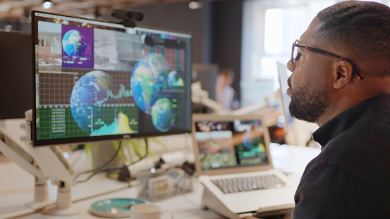 Stock photo of a black man, studying data on a screen. The data is associated with maps of the world, Earth.
