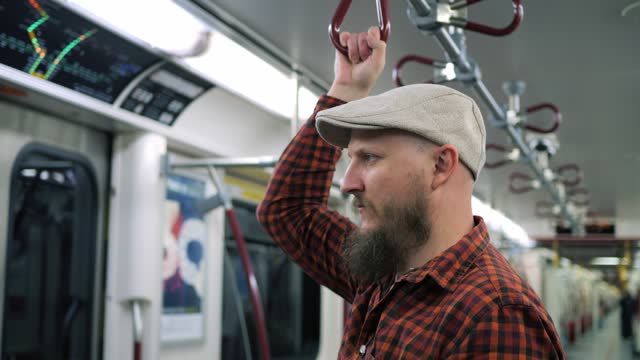 Thoughtful man in a cap holds handrail in subway. Close-up of bearded man in red checkered shirt goes on subway to downtown. Concept gloomy atmosphere in subway makes you stuck in your thoughts