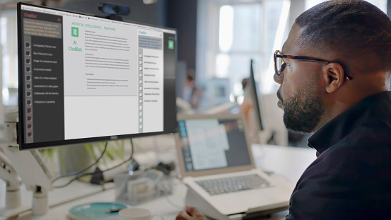 Stock image showing a black man’s face looking into a computer screen in an open plan working office. Type is being added to the screen by an Artificial intelligence,  AI,  chatbot.