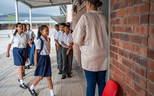 Woman teacher greeting elementary students while standing in classroom doorway