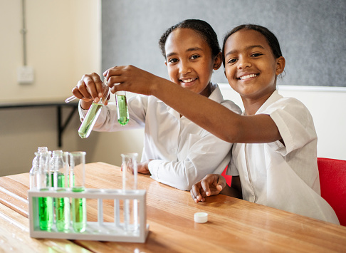 Two schoolgirls sitting at table in science class doing experiment with chemicals and smiling at camera