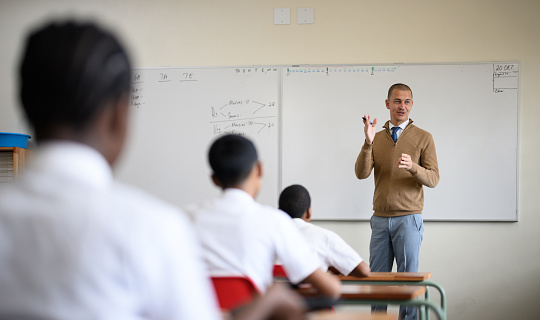 Over the shoulder view schoolchildren listen to teacher talking standing at the front of the classroom