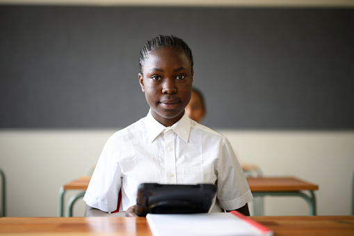 Portrait teenage African schoolgirl sitting at desk holding digital tablet