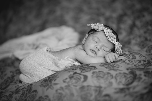Black and white photograph of a newborn girl who sleeps comfortably on her bed and wears a flower headband and a blanket