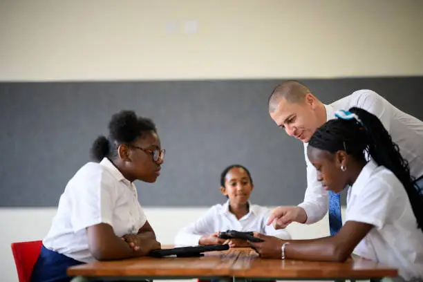 Photo of Male teacher helps schoolgirl with digital tablet sitting at desk