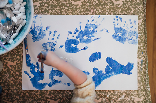 A high-angle, medium shot of an unrecognisable female toddler wearing casual clothing on a winter's day in Northumberland. She is at a preschool where she plays with different materials to develop her sensory perception. She does hand paintings on a piece of paper with blue paint.