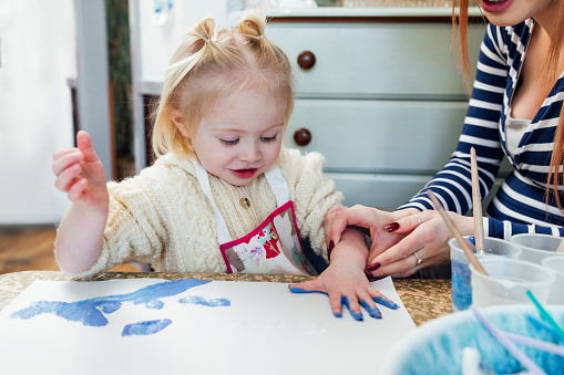 A medium shot of a female toddler and her mother wearing casual clothing on a winter's day in Northumberland. They are at a preschool where they sit at a table together and do a fingerpainting painting to develop the child's sensory perception.