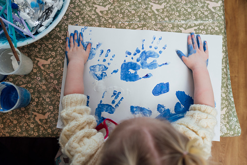 A high-angle, medium shot of an unrecognisable female toddler wearing casual clothing on a winter's day in Northumberland. She is at a preschool where she plays with different materials to develop her sensory perception. She does hand paintings on a piece of paper with blue paint.