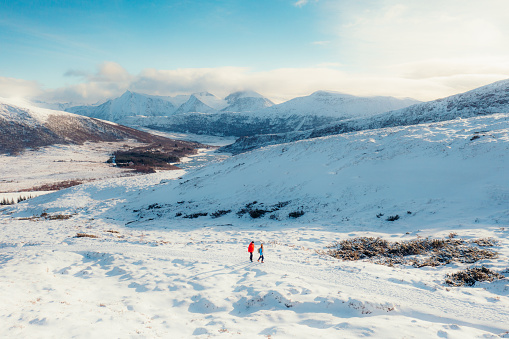 Drone panoramic photo of man in red and woman in blue down jackets contemplating hiking trip at the beautiful winter mountain landscape with a view of the fjord and beautiful lake during snowy blizzard and bright scenic sunset in the western fjords of Scandinavia