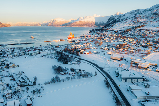 Drone panoramic photo of group of vehicles driving the highway in the beautiful Nordic city by the sea with boat deck and view of snowcapped mountain peaks during scenic colourful sunset in More og Romsdal, Scandinavia