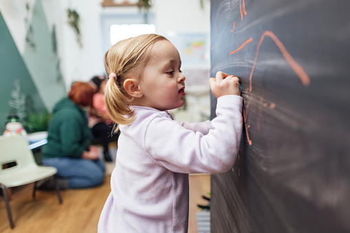 A side view, medium shot of a female toddler wearing casual clothing on a winter's day in Northumberland. She stands and draws on a chalkboard with coloured chalk in a creative hub for children in the North East of England.