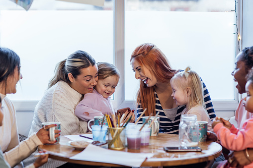 A medium shot of toddlers and their mothers wearing casual clothing on a winter's day in Northumberland. They are at a preschool where they sit around a table, talk and enjoy a cup of tea and snacks. One of the mothers shows her friends something on her smartphone and they laugh together.
