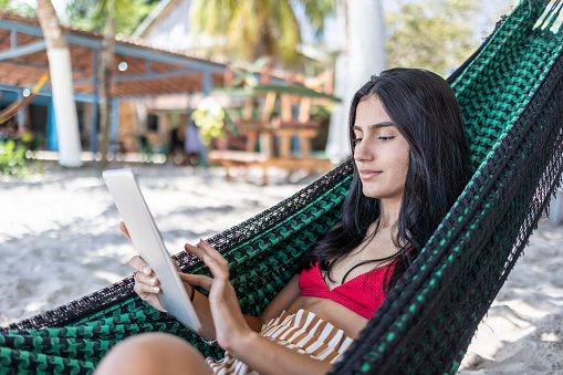 Latin woman in a hammock using a tablet on the beach