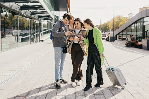 Two young women, one white and one of another ethnicity, embrace at a vibrant European railroad station, symbolizing the togetherness and joy of travel. They represent the generation Z and millennial generation, setting off on a backpacking adventure with smiles on their faces.