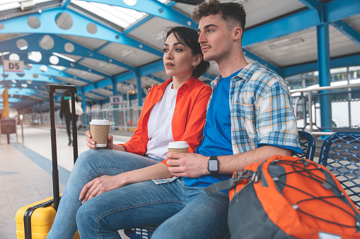 a young couple traveling together during their honeymoon, sitting in Interchange, waiting for bus, drinking coffee, chatting, reading news. Traveling together concept