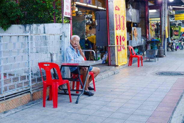 Thai senior man is sitting at small table on  sidewalk of street Wanghin Rd in Bangkok Ladprao. Man is sitting alone. He has gray hair and is wearing casual clothes. In background is a small restaurant with street street food and other local shops.