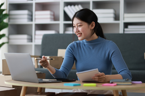 Asian woman starting a small business at home working with parcel boxes and calculating revenue and profit from sales.