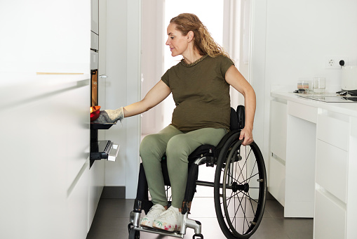 A woman in a wheelchair, at home in her kitchen, placing a cake into an adapted oven.