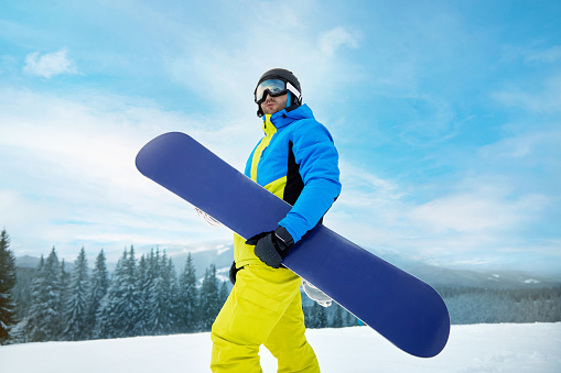 Portrait Of Man On The Background Of Mountains And Blue Sky, Holding Snowboard And Wearing Ski Glasses. Ski Goggles Of Snowboarder With The Reflection Of Snowed Mountains. Winter Sports