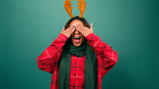 Excited young multi-ethnic woman covers eyes, Christmas reindeer studio. High quality photo