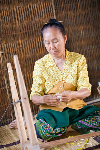 Thai mature woman is doing craft work with  loom outside of her farm house.  She is sitting on bamboo plaid in front of bamboo made wall. Woman is handling thread. Woman is wearing traditional clothing in green and yellow colors. Scene is  in village Parangmee in province Phitsanulok.
