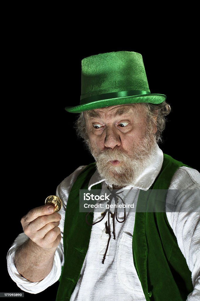 Irish leprechaun looks amazed at gold coin Classic Irish leprechaun with white beard, top hat, green velvet vest, looking amazed at a gold coin. He raises his eyebrows, purses lips and widens his eyes. Up lighting, isolated on black, vertical layout with copy space. Senior Adult Stock Photo