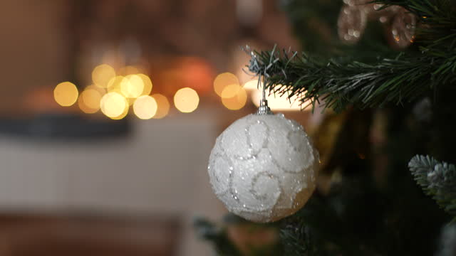 Closeup cropped shot of happy little girl hand hanging white festive ball on illuminated Christmas tree branches, on blurred background bright bokeh lights