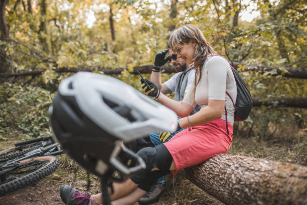 deux joyeux vététistes sont assis sur un tronc d’arbre dans la forêt et utilisent un téléphone portable - sportsman tree people recreational pursuit photos et images de collection