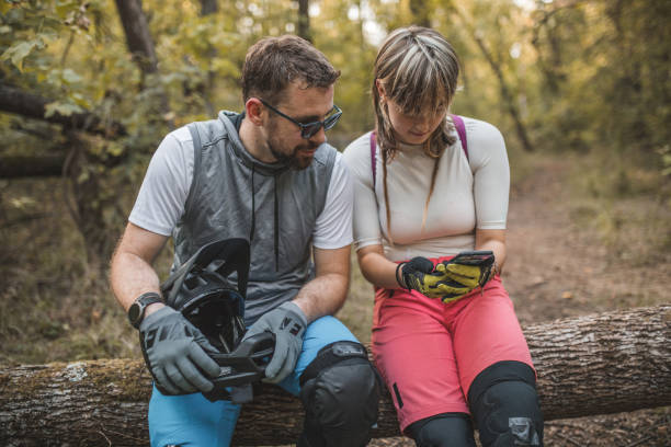 deux vététistes concentrés sont assis sur un tronc d’arbre dans la forêt, faisant une pause dans la descente de vtt et utilisant un téléphone portable - sportsman tree people recreational pursuit photos et images de collection