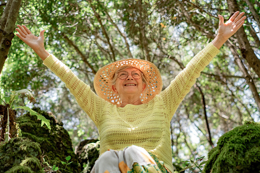 Smiling happy mature senior woman in outdoors excursion in the forest sitting with outstretched arms, elderly lady enjoying nature and freedom