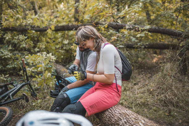deux vététistes sont assis sur un tronc d’arbre dans la forêt, faisant une pause dans la descente du vtt et utilisant un téléphone portable - sportsman tree people recreational pursuit photos et images de collection