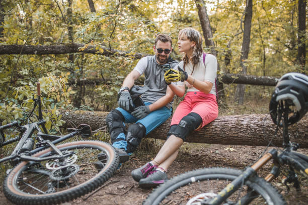 deux vététistes sont assis sur un tronc d’arbre dans la forêt, faisant une pause dans la descente et utilisant un téléphone portable pour la navigation - sportsman tree people recreational pursuit photos et images de collection