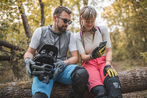 deux vététistes sont assis sur un tronc d’arbre, faisant une pause dans la descente et utilisant un téléphone portable - sportsman tree people recreational pursuit photos et images de collection