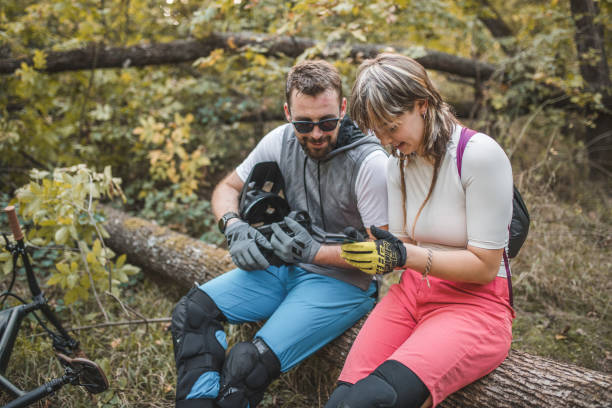 deux vététistes souriants sont assis sur un tronc d’arbre dans la forêt, faisant une pause dans la descente de vtt et utilisant un téléphone portable pour la navigation - sportsman tree people recreational pursuit photos et images de collection