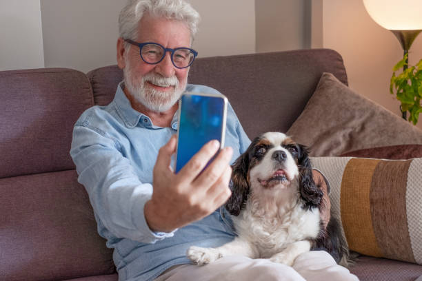 istock Senior smiling man sitting on home sofa using cellphone taking a selfie with his cavalier king Charles dog. Elderly bearded retired male and pet therapy concept 1771105091