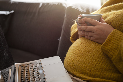 Pregnant woman sitting in the sofa with laptop in her lap and holding coffee mug. Concept of expecting woman working from home in the morning with sunlight. Photo taken in Sweden.