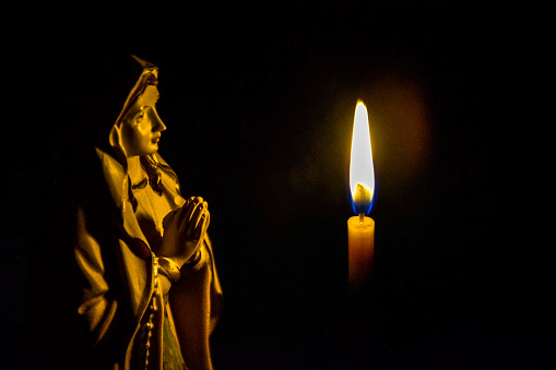 Christmas composition: statuette of the Virgin Mary and a lit church candle against dark background. Close-up. Selective focus. Copy space