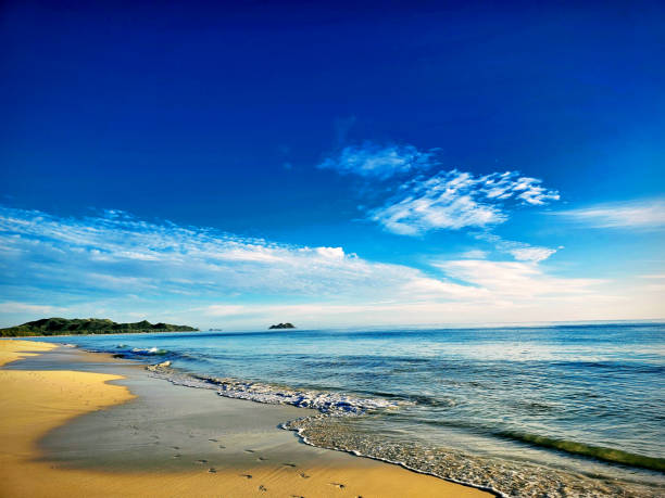 A scenic view of Waimanalo Beach, one of the longest uninterrupted white-sand beaches on Oahu, with golden sand and clear blue water, with green mountains and islands in the background. The image has a peaceful and serene mood. Waimanalo Beach is a historic and cultural site that may be one of the oldest inhabited settlements in Hawaii.