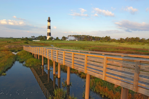 Wooden walkway from a viewpoint in the marsh leads to the Bodie Island lighthouse on the outer banks of North Carolina against a blue sky and white clouds