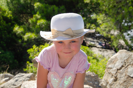 five-year-old girl in white hat smiles. She is wearing  pink t-shirt, green trees in background, blue eyes, blonde hair,