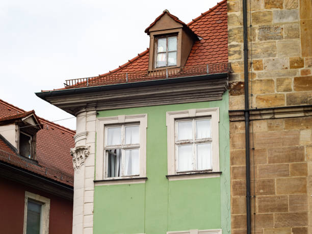 Narrow building next to a sandstone wall. Close up of the house exterior with the windows. The architecture is old and is part of the historical town center. On the rooftop is a gabled dormer.