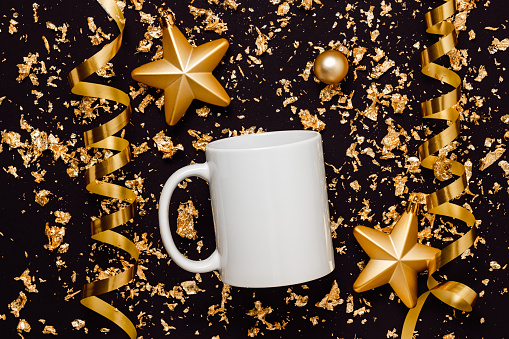 White mockup mug with christmas toys and golden ribbons on black background with golden glitter, top view. Mockup mug for logo, branding and design. Christmas, New Year and holidays concept